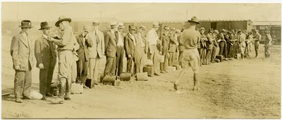 Fotografia orizzontale seppia che mostra un gruppo di uomini in abiti civili, molti dei quali hanno bagagli, in fila. Soldati in uniforme camminano lungo la fila parlando individualmente con gli uomini. Si vedono binari ferroviari e vagoni sullo sfondo da Carl Michel
